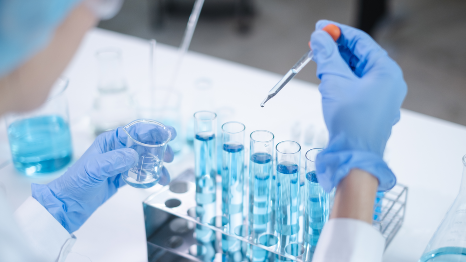 Лабораторная работа: рука с пипеткой добавляет жидкость в пробирки с голубой жидкостью. В другой руке держится мерный цилиндр. На столе расположены несколько пробирок и сосудов с реагентами.